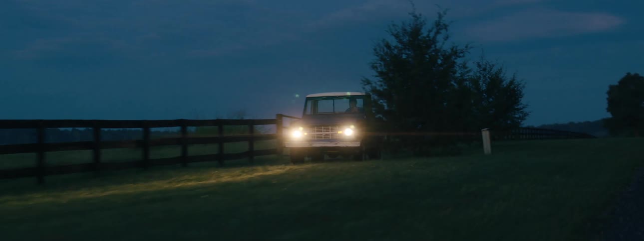 A vintage pickup truck drives through a field along a wooden fence