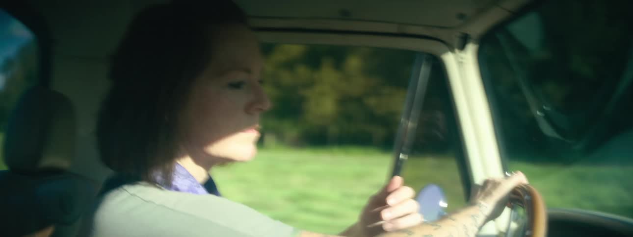 A woman driving a vehicle on a grassy field with trees the sky and clouds in the background