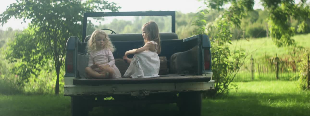 Sisters sit together in bed of truck parked in yard in countryside as elder combs youngers hair