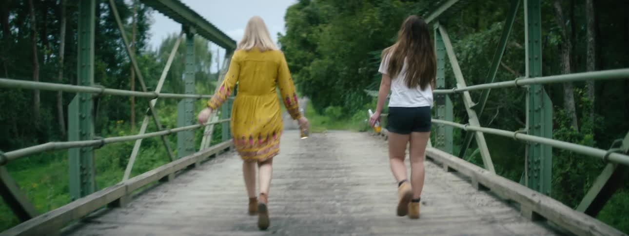A pair of women carrying half empty bottles of liquor walk together across a bridge on a cloudy day