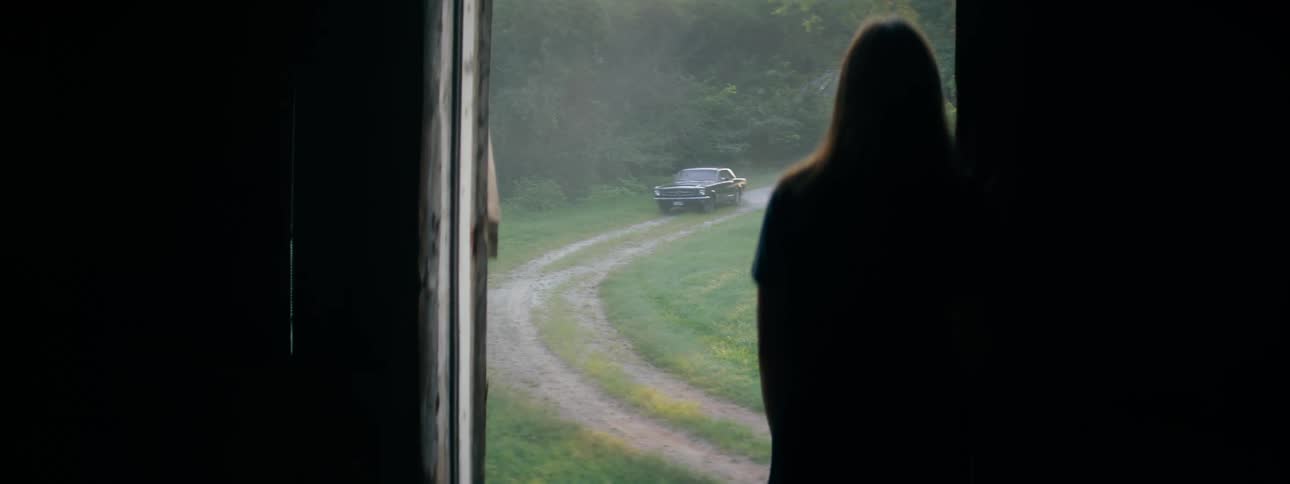 A silhouette of a woman standing in the doorway of a barn with a car driving on a track outside the doorway