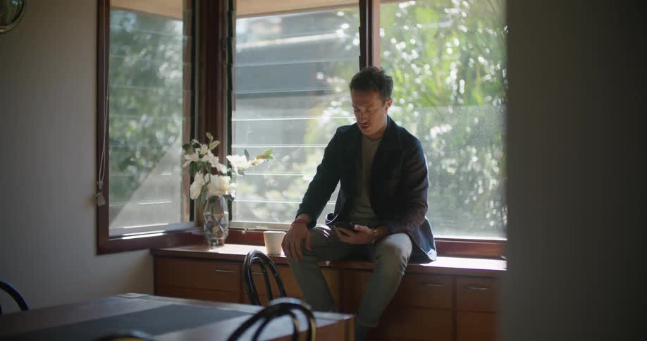 Man sits on furniture next to window talking on mobile phone