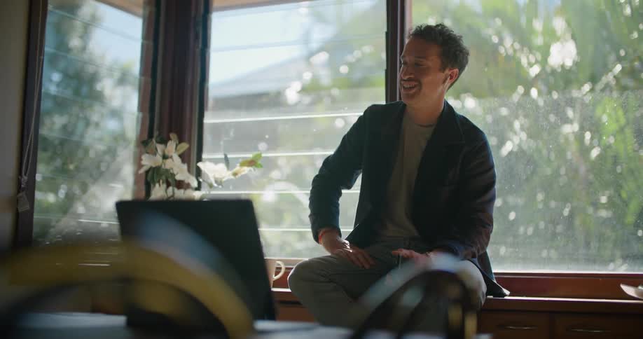 Man sits near window gesturing and talking on mobile phone across from laptop sitting on dining table