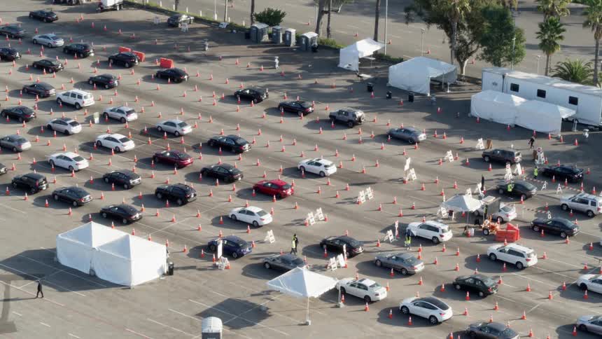 Rows of traffic travel slowly through lanes designated by traffic cones in a parking lot filled with tents and trailers