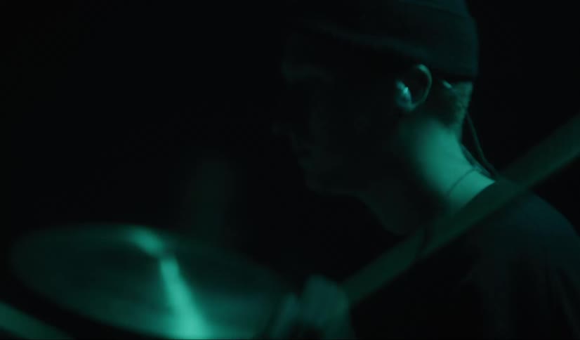 Drummer wearing earphones and knit hat plays drums on dark stage under light