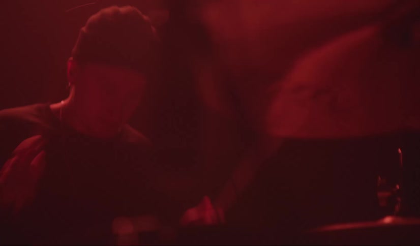 A man in a room sitting playing the drums with drumsticks with a wall in the background
