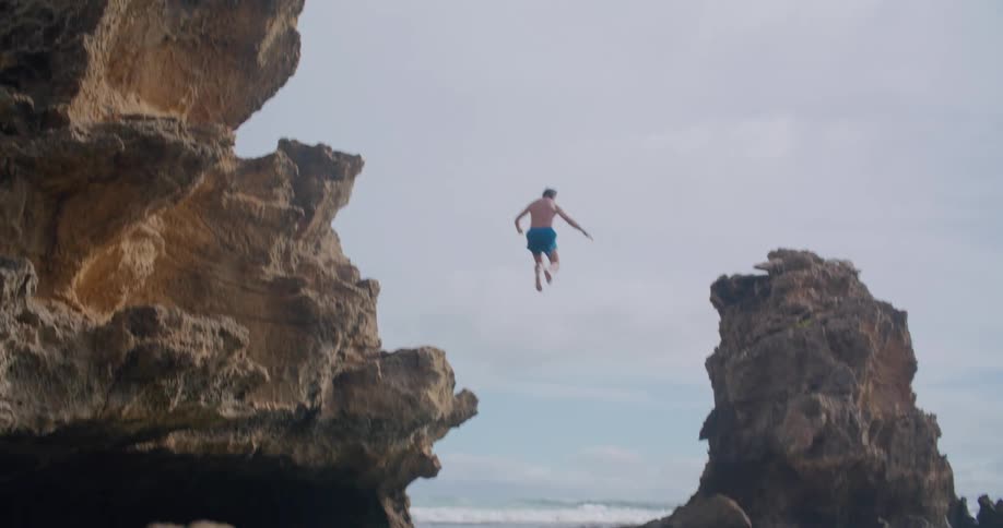 A man high fives his friend then jumps off the cliff and splashes into the ocean