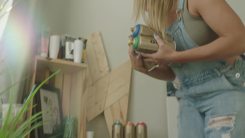 Blonde artist adds another spray paint can to those she is holding as she stands and walks past wall of art