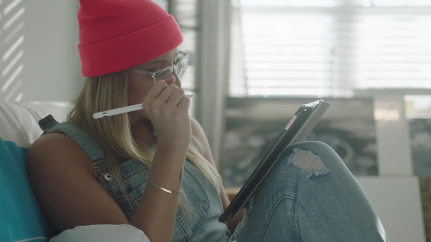 Blonde woman sits on couch sketching on tablet with paintings leaning on wall under window