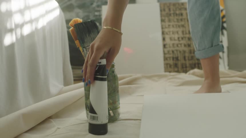 Woman places a tube of paint on the floor next to a jar holding paint brushes