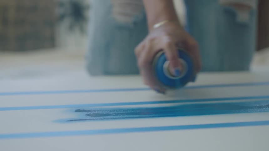 A kneeling woman uses spray paint to draw on a board lying on the floor