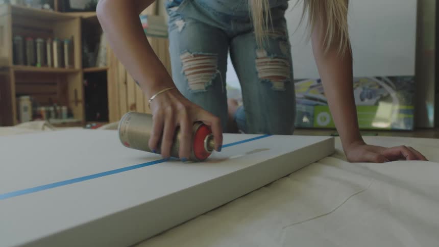 A woman shakes a spray paint can as she paints on a taped canvas on a tarp