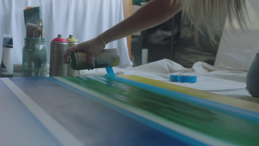 Kneeling blonde female artist spray paints green onto canvas