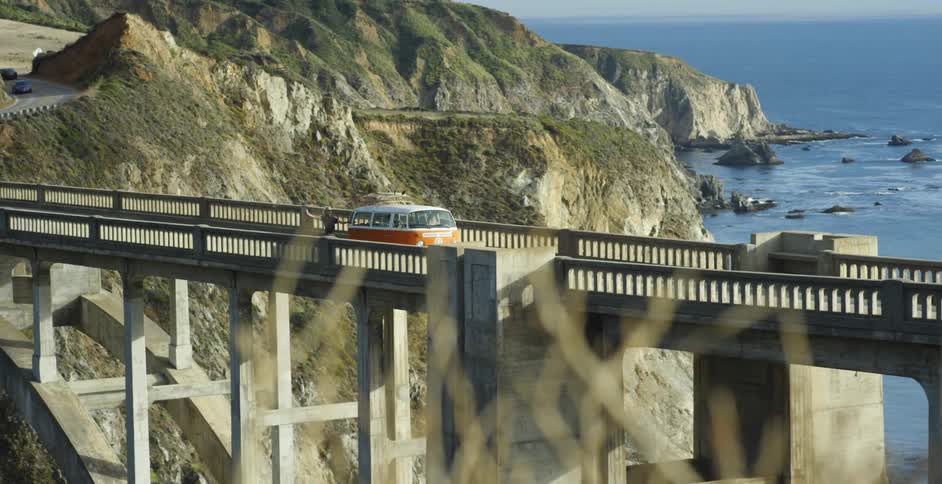 A v.w. bus towing a skateboarder is driving on a bridge along the coast in daylight
