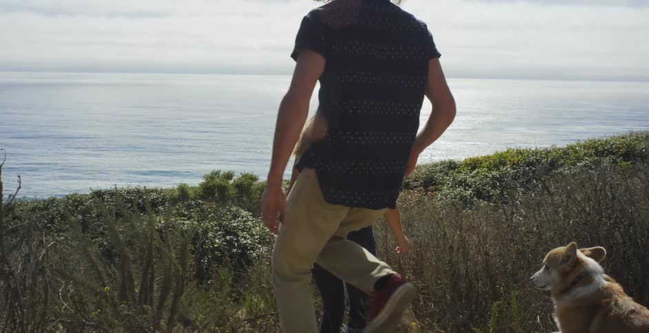 A couple and their corgi are walking down a hillside toward the ocean on a sunny day