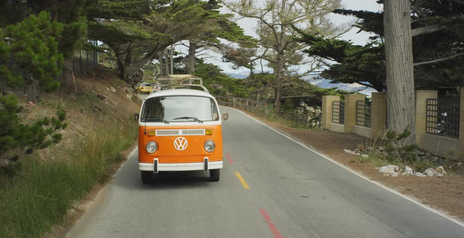 A v.w. bus is driving on a rural road along the coast in daylight