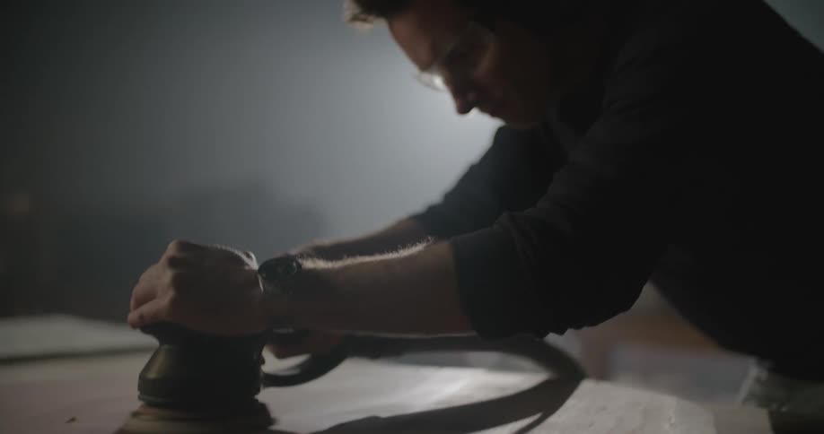 A man in a workshop with goggles and earmuffs using an electric sander with a light in the background