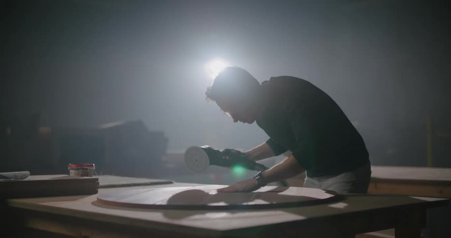 A carpenter users a sander on a round piece of wood and leans down to tap the surface