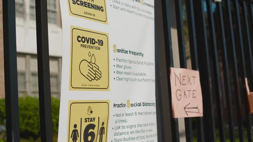 Covid 19 poster sits on gate near hand written sign as another torn sign flutters in the breeze