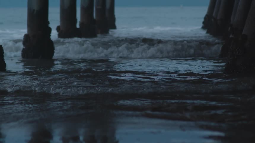 A beach underneath a pier with pillars with the ocean sky and clouds in the background