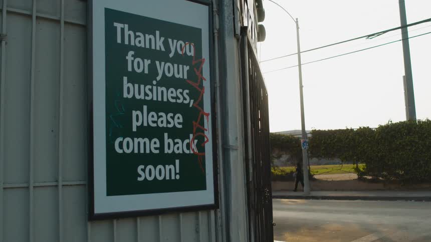 A pedestrian and a car pass by a business with a sign hanging on it