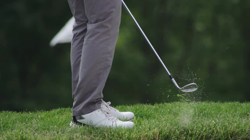A person hitting a golf ball with a golf club on grass with a flag and trees in the background