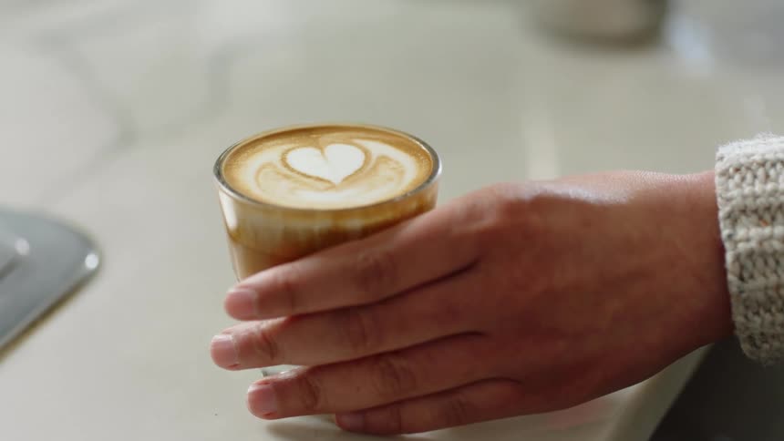 A person in a room placing a glass of cappuccino on a table with a heart design in the foam