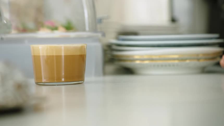Latte in short glass sits on white surface near glass dome covered food and gold rimmed plates