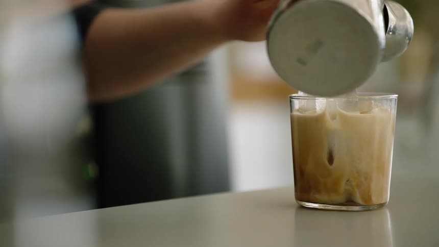 A person in a room pouring coffee and cream into a glass with ice on a table