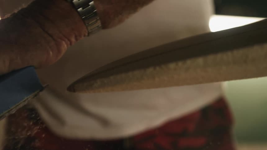 A man is sanding a wooden surfboard with a sanding block