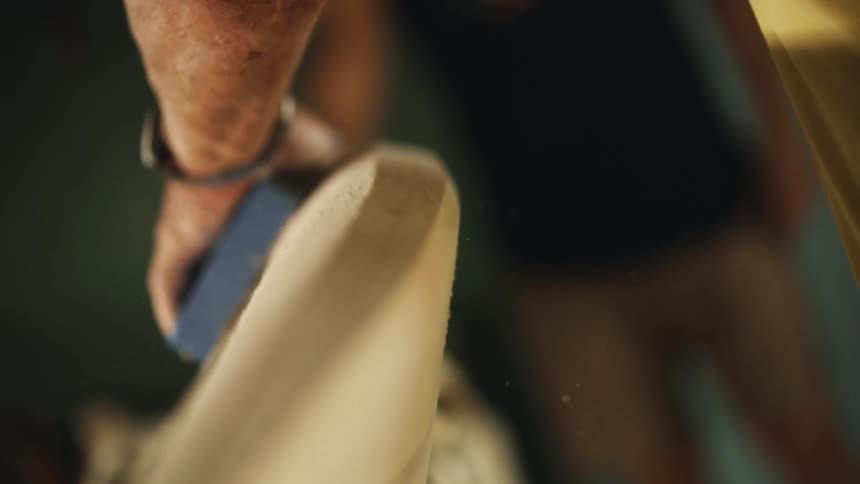 A man sands an unpainted surfboard with a sanding pad