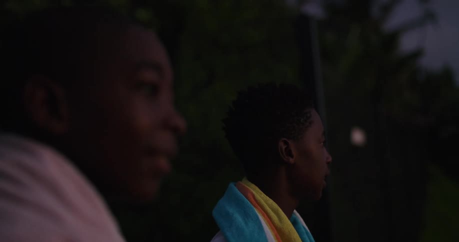 A pair of boys with towels around their shoulders talk with one another while standing outdoors in the evening