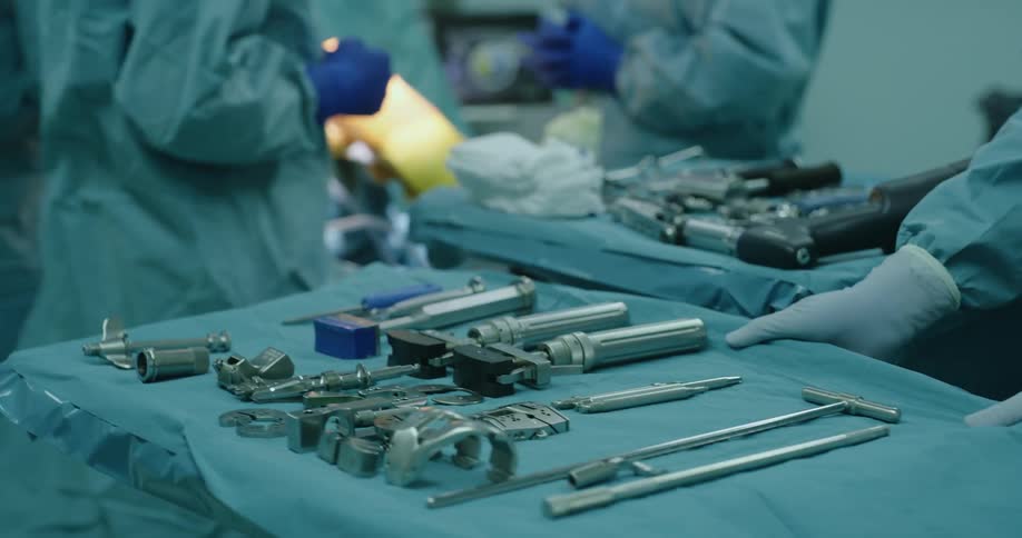Surgical assistant rolls in cart with medical tools toward surgical team standing near patient
