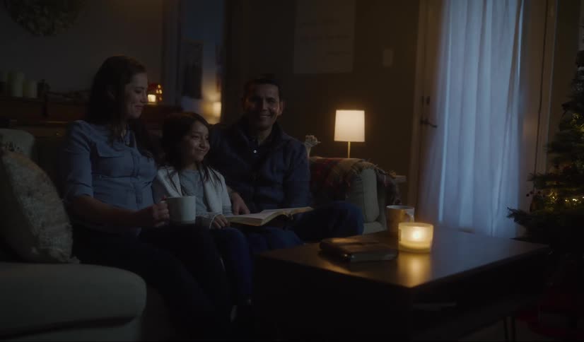Mom and dad sit on a couch with their daughter between then smiling and happy in a room lit by candles and christmas tree
