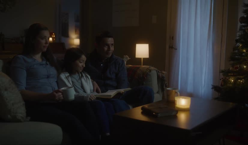 A man a woman and a girl in a room sitting on a couch next to a table with a christmas tree in the background