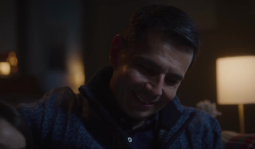 A father sits on a couch at home with his daughter and looks down at her and they smile at one another