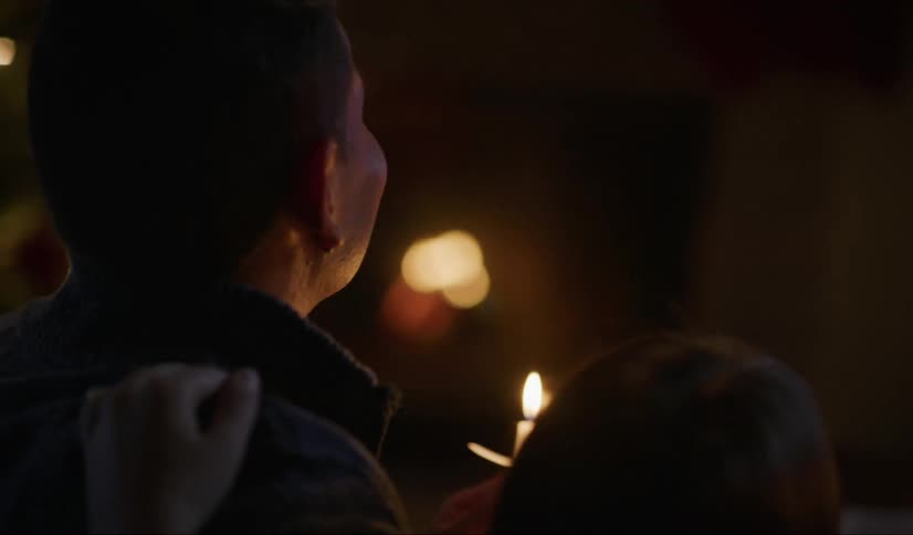 A man holding a flaming candle turns and leans his head on woman beside him