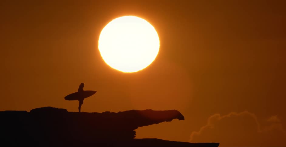 A surfer stands on a cliff at sunrise with his surfboard under his arm