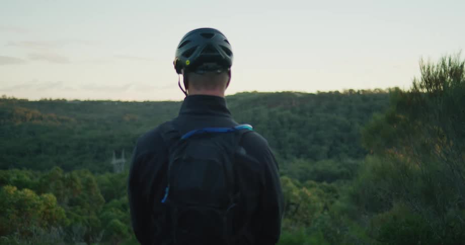 Man wearing bicycle helmet stands over looking hills and trees surrounding structure