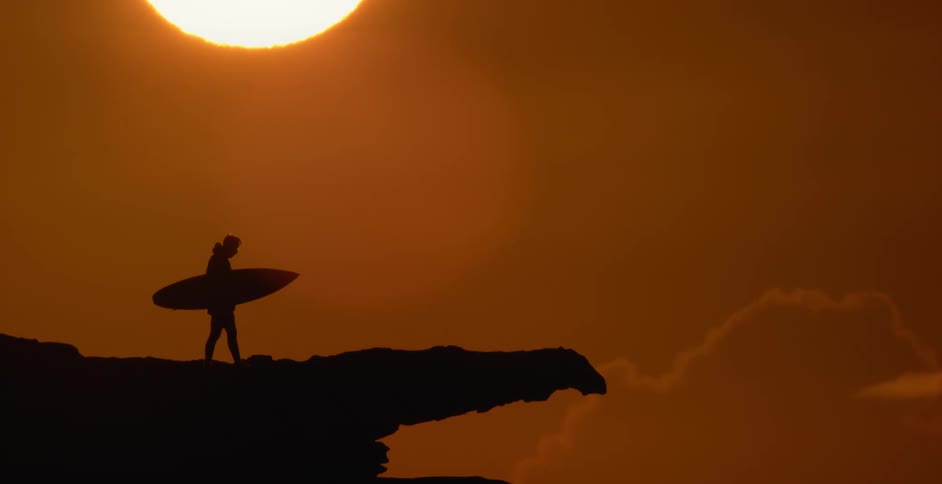 Silhouette of a man carrying a surfboard on a promontory at sunset
