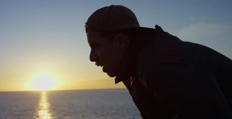 A man with a hat standing near the coast with the ocean the sunset the sky and clouds in the background