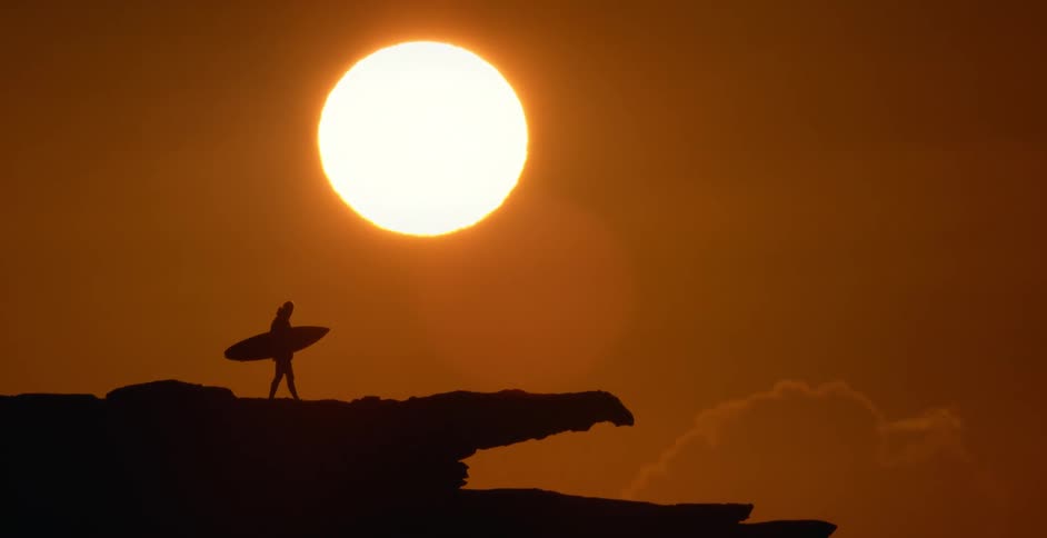 Silhouette of a person walking on a rocky ledge holding a surfboard with a bright sun in a red sky