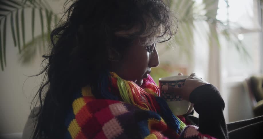 A woman sits wrapped in a blanket and sipping on a mug of a hot drink