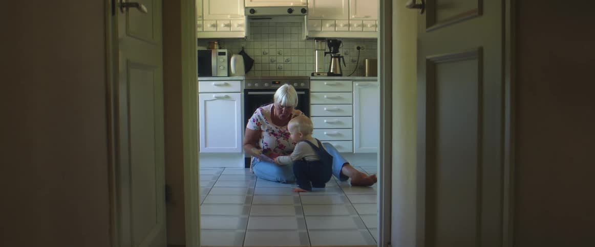 A elderly woman sits on the kitchen floor while showing a story book to a small child