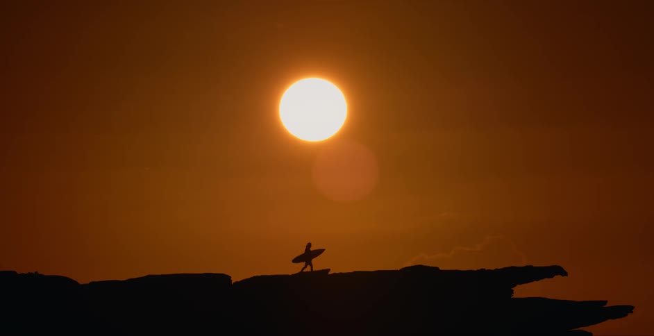 Silhouette of a person standing on a rocky ledge holding a surfboard under a bright sun in a red sky