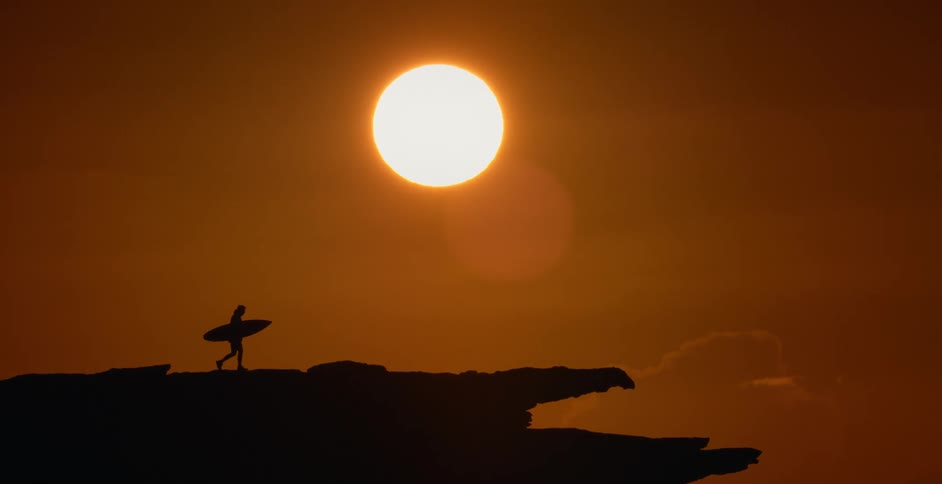 A silhouette of a person walking with a surfboard with the sun the sky and clouds in the background