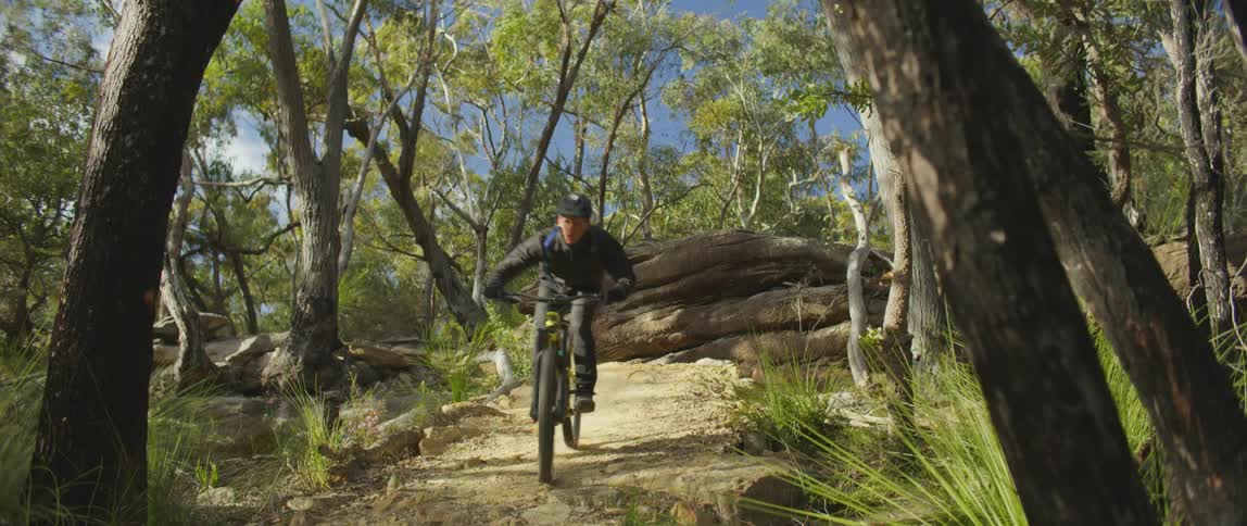 Man wearing helmet rides mountain bike along trail through woods taking jump