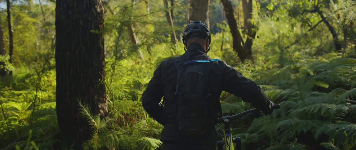 A person with a helmet and backpack walking a bicycle in a forest with the sky and clouds in the background
