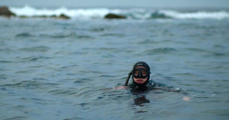 Man wearing snorkel and wet suit threads water in ocean as waves hit rocks in background
