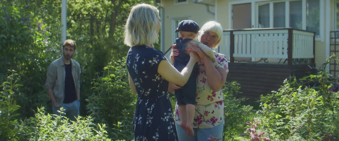 A family is outside of a home and the mother hands the infant to the grandmother